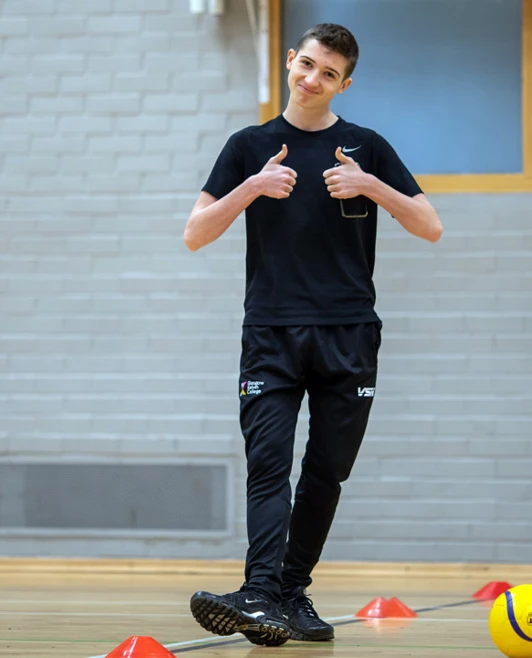 Smiling student giving two thumbs up while standing on a gym hall floor near cones and a ball. Smiling student giving two thumbs up while standing on a gym hall floor near cones and a ball.